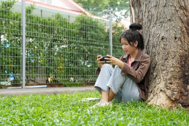 Bir lise öğrencisi bir ağaca yaslanmış, akıllı telefonuyla oyun oynarken gülümsüyor, odaklanmış ve eğlenmiş görünüyor..