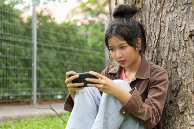 Bir lise öğrencisi bir ağaca yaslanmış, akıllı telefonuyla oyun oynarken gülümsüyor, odaklanmış ve eğlenmiş görünüyor..