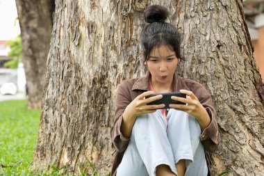 Bir lise öğrencisi bir ağaca yaslanmış, akıllı telefonuyla oyun oynarken gülümsüyor, odaklanmış ve eğlenmiş görünüyor..