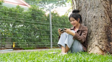 Bir lise öğrencisi bir ağaca yaslanmış, akıllı telefonuyla oyun oynarken gülümsüyor, odaklanmış ve eğlenmiş görünüyor..