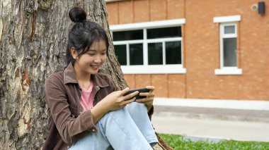 Bir lise öğrencisi bir ağaca yaslanmış, akıllı telefonuyla oyun oynarken gülümsüyor, odaklanmış ve eğlenmiş görünüyor..