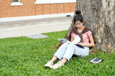 Bir lise öğrencisi, derslerine çok dalmış bir şekilde bir ağaca karşı oturuyor. Elinde bir kitap tutarken bir deftere yazmaya odaklanmış..