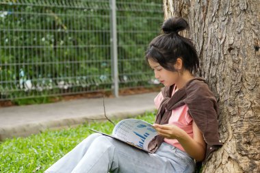 Bir lise öğrencisi, derslerine çok dalmış bir şekilde bir ağaca karşı oturuyor. Elinde bir kitap tutarken bir deftere yazmaya odaklanmış..