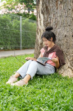 Bir lise öğrencisi, derslerine çok dalmış bir şekilde bir ağaca karşı oturuyor. Elinde bir kitap tutarken bir deftere yazmaya odaklanmış..