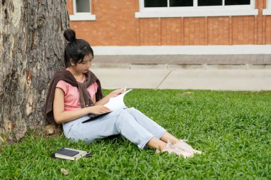 Bir lise öğrencisi, derslerine çok dalmış bir şekilde bir ağaca karşı oturuyor. Elinde bir kitap tutarken bir deftere yazmaya odaklanmış..