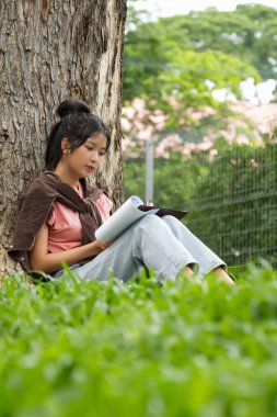 Bir lise öğrencisi, derslerine çok dalmış bir şekilde bir ağaca karşı oturuyor. Elinde bir kitap tutarken bir deftere yazmaya odaklanmış..