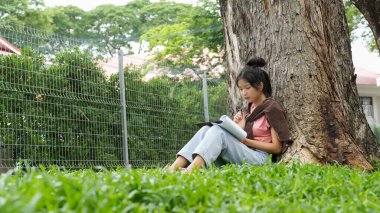 Bir lise öğrencisi, derslerine çok dalmış bir şekilde bir ağaca karşı oturuyor. Elinde bir kitap tutarken bir deftere yazmaya odaklanmış..