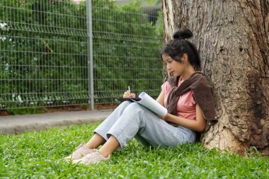Bir lise öğrencisi, derslerine çok dalmış bir şekilde bir ağaca karşı oturuyor. Elinde bir kitap tutarken bir deftere yazmaya odaklanmış..