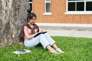 Bir lise öğrencisi tabletine yazmaya odaklanmış bir şekilde bir ağaca yaslanır. Okulun kırmızı tuğla dış görünüşü arka planda görünüyor..