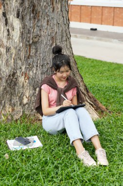Bir lise öğrencisi tabletine yazmaya odaklanmış bir şekilde bir ağaca yaslanır. Okulun kırmızı tuğla dış görünüşü arka planda görünüyor..