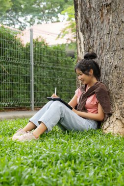 Bir lise öğrencisi tabletine yazmaya odaklanmış bir şekilde bir ağaca yaslanır. Okulun kırmızı tuğla dış görünüşü arka planda görünüyor..