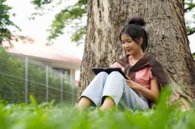 Bir lise öğrencisi tabletine yazmaya odaklanmış bir şekilde bir ağaca yaslanır. Okulun kırmızı tuğla dış görünüşü arka planda görünüyor..