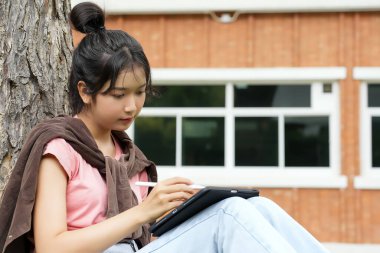 Bir lise öğrencisi tabletine yazmaya odaklanmış bir şekilde bir ağaca yaslanır. Okulun kırmızı tuğla dış görünüşü arka planda görünüyor..