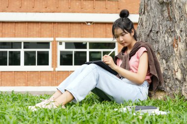 Bir lise öğrencisi tabletine yazmaya odaklanmış bir şekilde bir ağaca yaslanır. Okulun kırmızı tuğla dış görünüşü arka planda görünüyor..