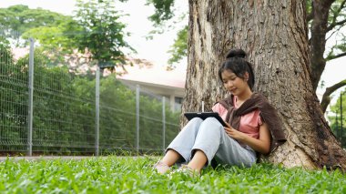Bir lise öğrencisi tabletine yazmaya odaklanmış bir şekilde bir ağaca yaslanır. Okulun kırmızı tuğla dış görünüşü arka planda görünüyor..