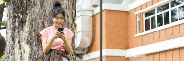 Bir lise öğrencisi telefonuna bakarken gülümseyen bir ağaca yaslanır. Okulun kırmızı tuğla dış görünüşü arka planda görünüyor..