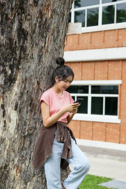 Bir lise öğrencisi telefonuna bakarken gülümseyen bir ağaca yaslanır. Okulun kırmızı tuğla dış görünüşü arka planda görünüyor..