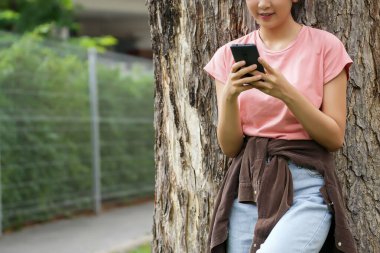Bir lise öğrencisi telefonuna bakarken gülümseyen bir ağaca yaslanır. Okulun kırmızı tuğla dış görünüşü arka planda görünüyor..