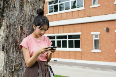 Bir lise öğrencisi bir ağaca karşı oturuyor, akıllı telefonuyla oyun oynarken gülümsüyor. Arka plan okulun kırmızı tuğla dış görünüşünü gösteriyor, rahatlığı arttırıyor.