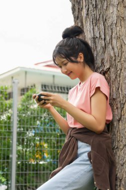 Bir lise öğrencisi bir ağaca karşı oturuyor, akıllı telefonuyla oyun oynarken gülümsüyor. Arka plan okulun kırmızı tuğla dış görünüşünü gösteriyor, rahatlığı arttırıyor.