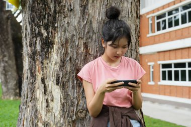 Bir lise öğrencisi bir ağaca karşı oturuyor, akıllı telefonuyla oyun oynarken gülümsüyor. Arka plan okulun kırmızı tuğla dış görünüşünü gösteriyor, rahatlığı arttırıyor.