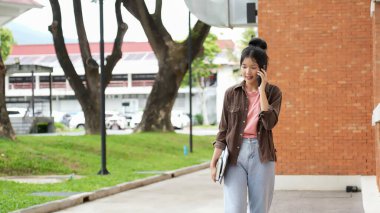 Bir lise öğrencisi kırmızı tuğlalı okul binasında yürüyor ve telefonuyla konuşuyor. Diğer elinde kitap ve tablet tutuyor..