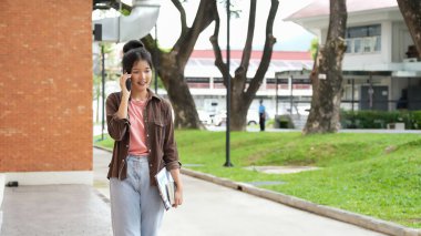 Bir lise öğrencisi kırmızı tuğlalı okul binasında yürüyor ve telefonuyla konuşuyor. Diğer elinde kitap ve tablet tutuyor..
