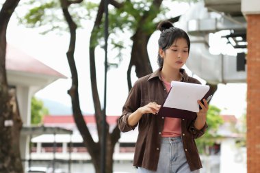 Bir lise öğrencisi kırmızı tuğla bir duvara yaslanmış kitap okuyor. Pembe gömlek, açık mavi kot pantolon ve kahverengi ceket giyiyor. O kitabına odaklanmış, hafifçe gülümsüyor..