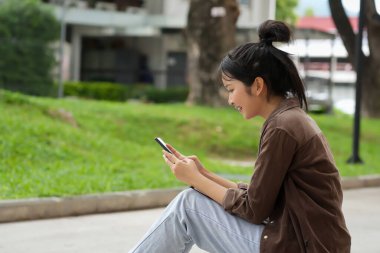 Bir lise öğrencisi kırmızı tuğla bir duvara karşı kaldırımda oturuyor ve telefonuna bakıyor. Pembe gömlek, açık mavi kot pantolon ve kahverengi ceket giyiyor. Kendini telefonuna kaptırmış..