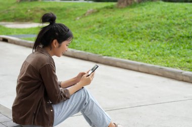Bir lise öğrencisi kırmızı tuğla bir duvara karşı kaldırımda oturuyor ve telefonuna bakıyor. Pembe gömlek, açık mavi kot pantolon ve kahverengi ceket giyiyor. Kendini telefonuna kaptırmış..
