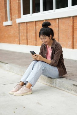 Bir lise öğrencisi kırmızı tuğla bir duvara karşı kaldırımda oturuyor ve telefonuna bakıyor. Pembe gömlek, açık mavi kot pantolon ve kahverengi ceket giyiyor. Kendini telefonuna kaptırmış..