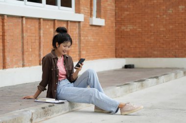 Bir lise öğrencisi kırmızı tuğla bir duvara karşı kaldırımda oturuyor ve telefonuna bakıyor. Pembe gömlek, açık mavi kot pantolon ve kahverengi ceket giyiyor. Kendini telefonuna kaptırmış..