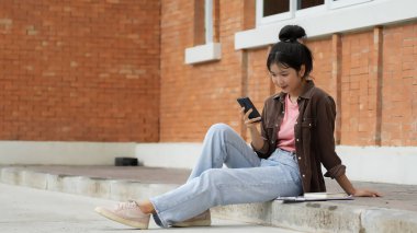 Bir lise öğrencisi kırmızı tuğla bir duvara karşı kaldırımda oturuyor ve telefonuna bakıyor. Pembe gömlek, açık mavi kot pantolon ve kahverengi ceket giyiyor. Kendini telefonuna kaptırmış..