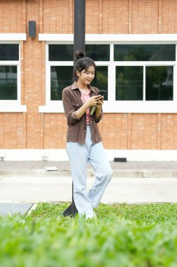 Bir lise öğrencisi okul binasının önünde telefonuna bakarak yürüyor. Gülümsüyor ve telefonuyla meşgul görünüyor..