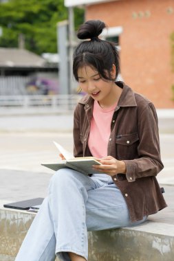 Bir lise öğrencisi okul binasının önünde telefonuna bakarak yürüyor. Gülümsüyor ve telefonuyla meşgul görünüyor.