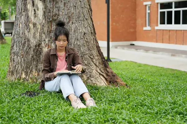 Modern bir okul binasının önünde oturan bir lise öğrencisi deftere bir şeyler yazıyor. Kahverengi ceket ve açık mavi kot giyiyor. Yazılarına odaklanmış.. 