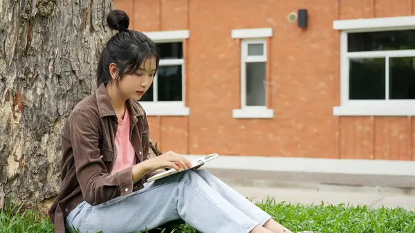 Modern bir okul binasının önünde oturan bir lise öğrencisi deftere bir şeyler yazıyor. Kahverengi ceket ve açık mavi kot giyiyor. Yazılarına odaklanmış.. 
