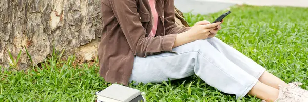 Bir lise öğrencisi bir ağacın altındaki çimlerin üzerinde oturuyor, akıllı telefonuna bakarken gülümsüyor. Kahverengi bir ceket ve açık mavi kot pantolon giyiyor, rahat ve mutlu görünüyor.. 
