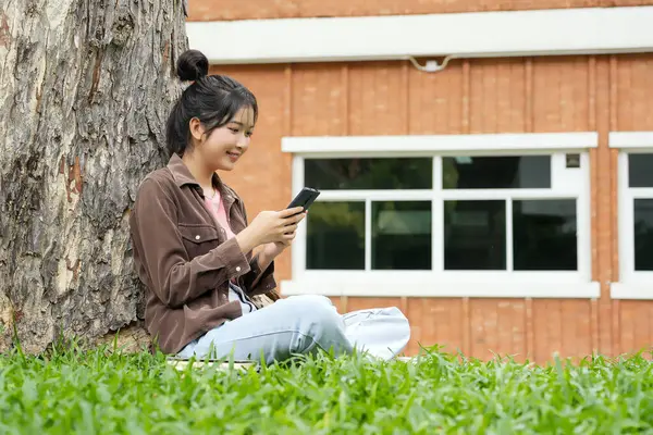 Bir lise öğrencisi bir ağacın altındaki çimlerin üzerinde oturuyor, akıllı telefonuna bakarken gülümsüyor. Kahverengi bir ceket ve açık mavi kot pantolon giyiyor, rahat ve mutlu görünüyor.. 