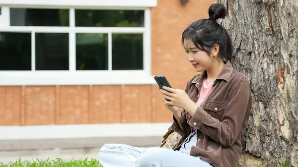Bir lise öğrencisi bir ağacın altındaki çimlerin üzerinde oturuyor, akıllı telefonuna bakarken gülümsüyor. Kahverengi bir ceket ve açık mavi kot pantolon giyiyor, rahat ve mutlu görünüyor.. 