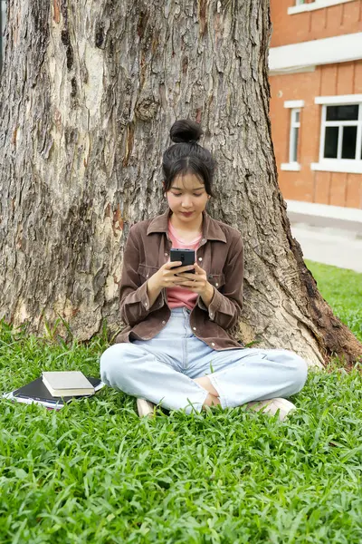 Bir lise öğrencisi bir ağacın altındaki çimlerin üzerinde oturuyor, akıllı telefonuna bakarken gülümsüyor. Kahverengi bir ceket ve açık mavi kot pantolon giyiyor, rahat ve mutlu görünüyor.. 