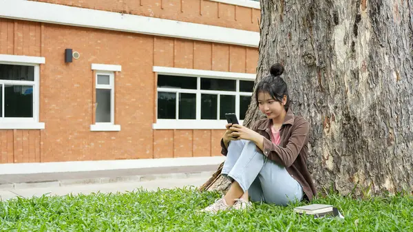 Bir lise öğrencisi bir ağacın altındaki çimlerin üzerinde oturuyor, akıllı telefonuna bakarken gülümsüyor. Kahverengi bir ceket ve açık mavi kot pantolon giyiyor, rahat ve mutlu görünüyor.. 