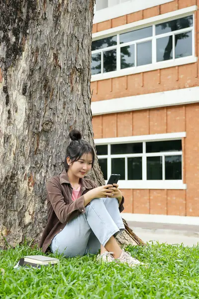 Bir lise öğrencisi bir ağacın altındaki çimlerin üzerinde oturuyor, akıllı telefonuna bakarken gülümsüyor. Kahverengi bir ceket ve açık mavi kot pantolon giyiyor, rahat ve mutlu görünüyor.. 