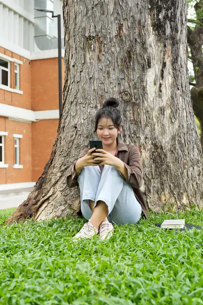 Bir lise öğrencisi bir ağacın altındaki çimlerin üzerinde oturuyor, akıllı telefonuna bakarken gülümsüyor. Kahverengi bir ceket ve açık mavi kot pantolon giyiyor, rahat ve mutlu görünüyor.. 