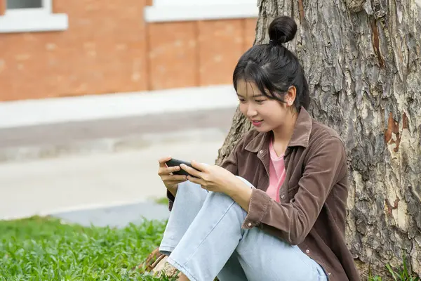 Bir lise öğrencisi bir ağaca yaslanmış, akıllı telefonuyla oyun oynarken gülümsüyor, odaklanmış ve eğlenmiş görünüyor..