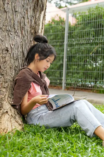 Bir lise öğrencisi, derslerine çok dalmış bir şekilde bir ağaca karşı oturuyor. Elinde bir kitap tutarken bir deftere yazmaya odaklanmış..