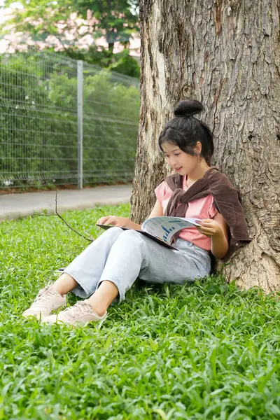Bir lise öğrencisi, derslerine çok dalmış bir şekilde bir ağaca karşı oturuyor. Elinde bir kitap tutarken bir deftere yazmaya odaklanmış..