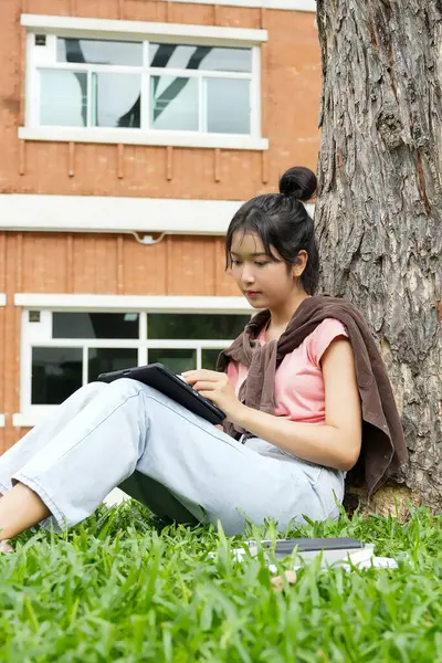 Bir lise öğrencisi tabletine yazmaya odaklanmış bir şekilde bir ağaca yaslanır. Okulun kırmızı tuğla dış görünüşü arka planda görünüyor..