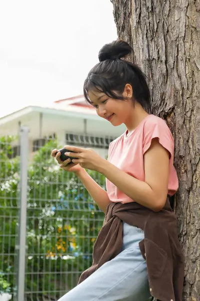 Bir lise öğrencisi bir ağaca karşı oturuyor, akıllı telefonuyla oyun oynarken gülümsüyor. Arka plan okulun kırmızı tuğla dış görünüşünü gösteriyor, rahatlığı arttırıyor.