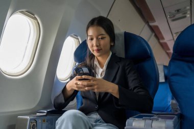 İş kadını mesajlaşmak için akıllı telefonu kullanıyor ve tatil zamanı yaşam tarzı sırasında uçuştaki iş seyahatlerinde pencerelerin yanında otururken finans sektörü raporlarını okumak için çalışıyor..